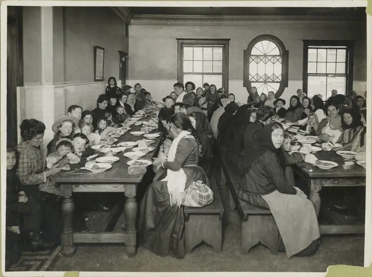 early 1900s photograph of immigrants eating at long wooden tables at Ellis Island.  Many of the immigrants are dressed in the style of their home country, with women wearing head coverings.  There are many children present along with the women.